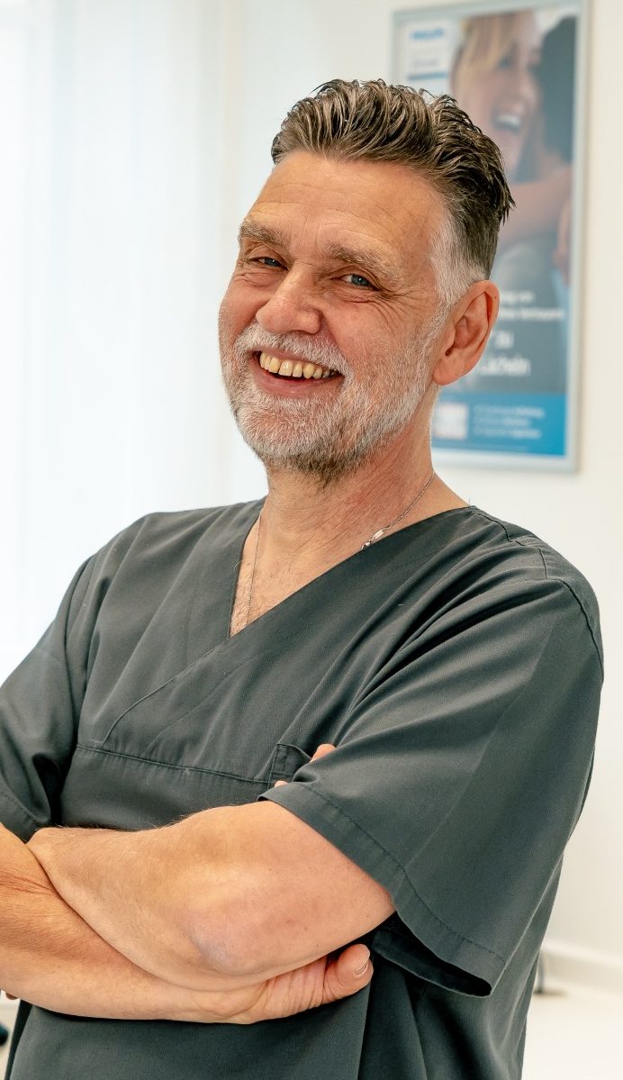 Dr. med. dent. Michael Fuchs Lächelnder Arzt in grauer Arbeitskleidung mit verschränkten Armen.
