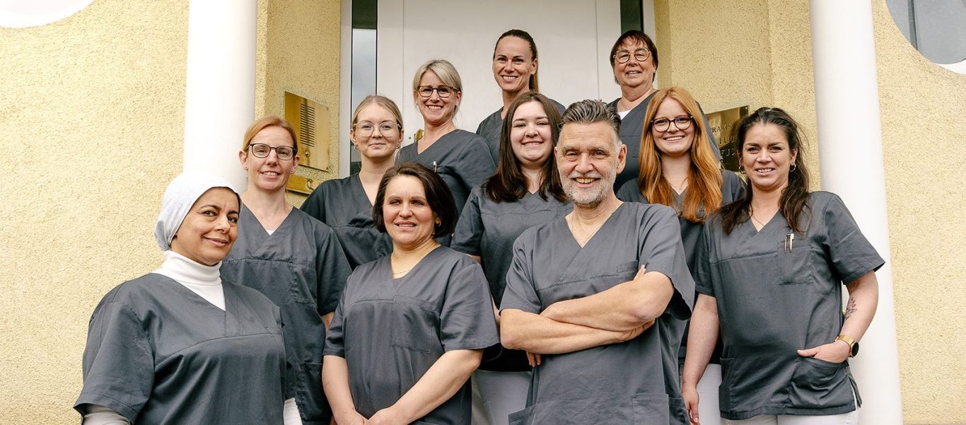 Unser Praxisteam Gruppe von medizinischem Personal in graueren Arbeitskleidung vor einer Tür.
