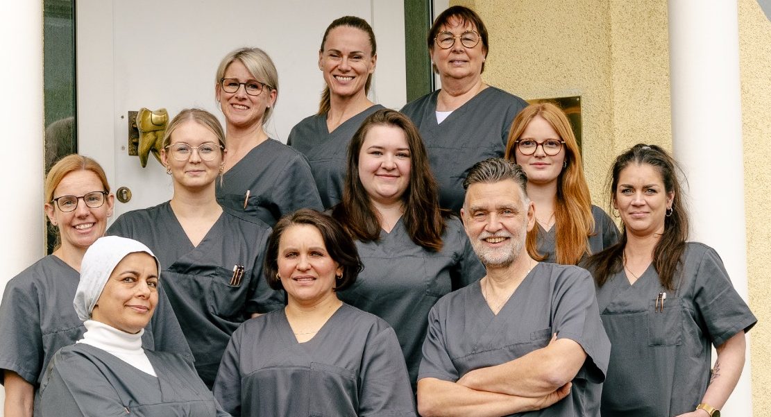 Unser Praxisteam Gruppe von medizinischem Personal in graueren Arbeitskleidung vor einer Tür.