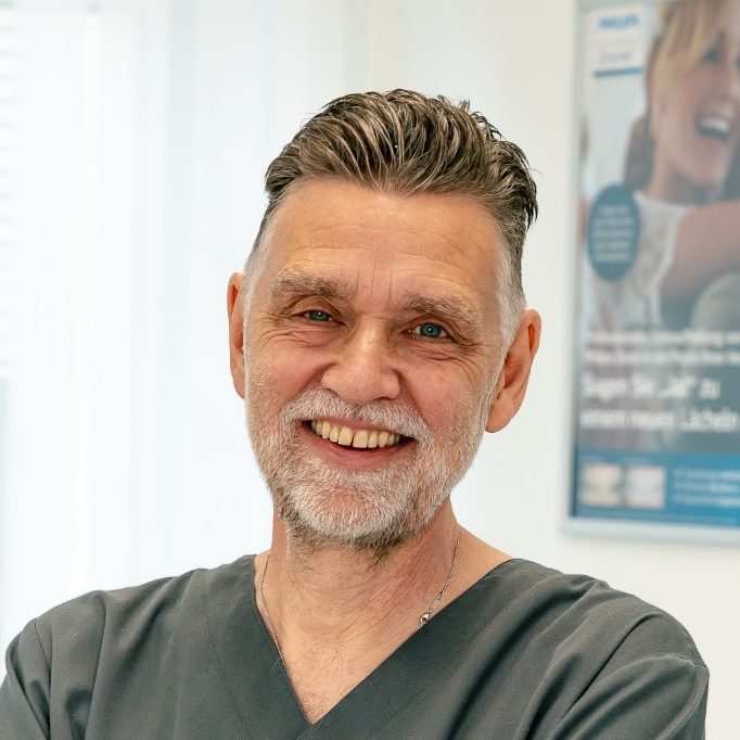 Dr. med. dent. Michael Fuchs Portrait eines lächelnden Mannes in einem Arztkittel, mit fröhlichem Hintergrund.