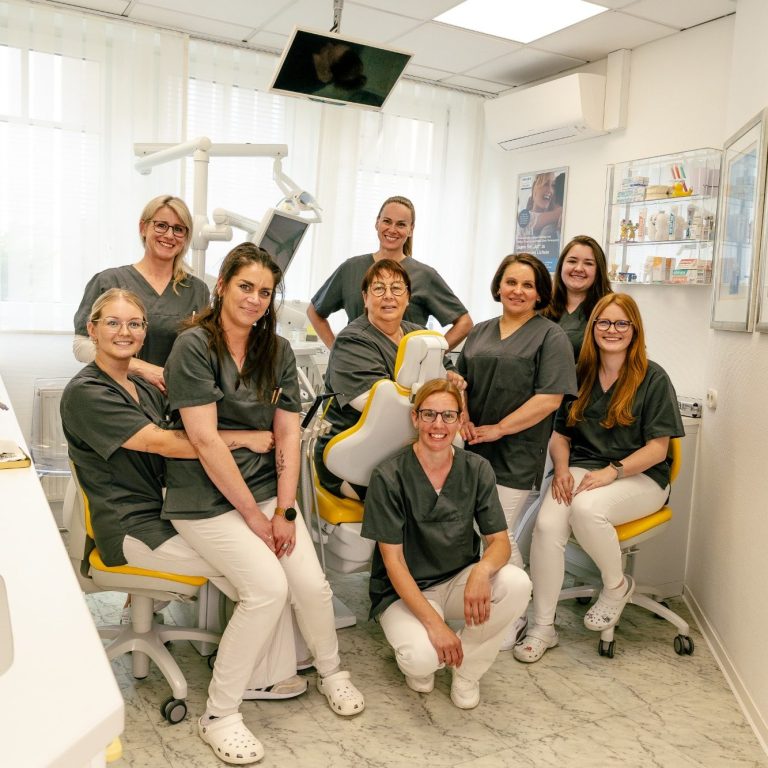 Ein Team von zehn Dentalmitarbeitern in grauen Uniformen lächelt in einer Praxis.